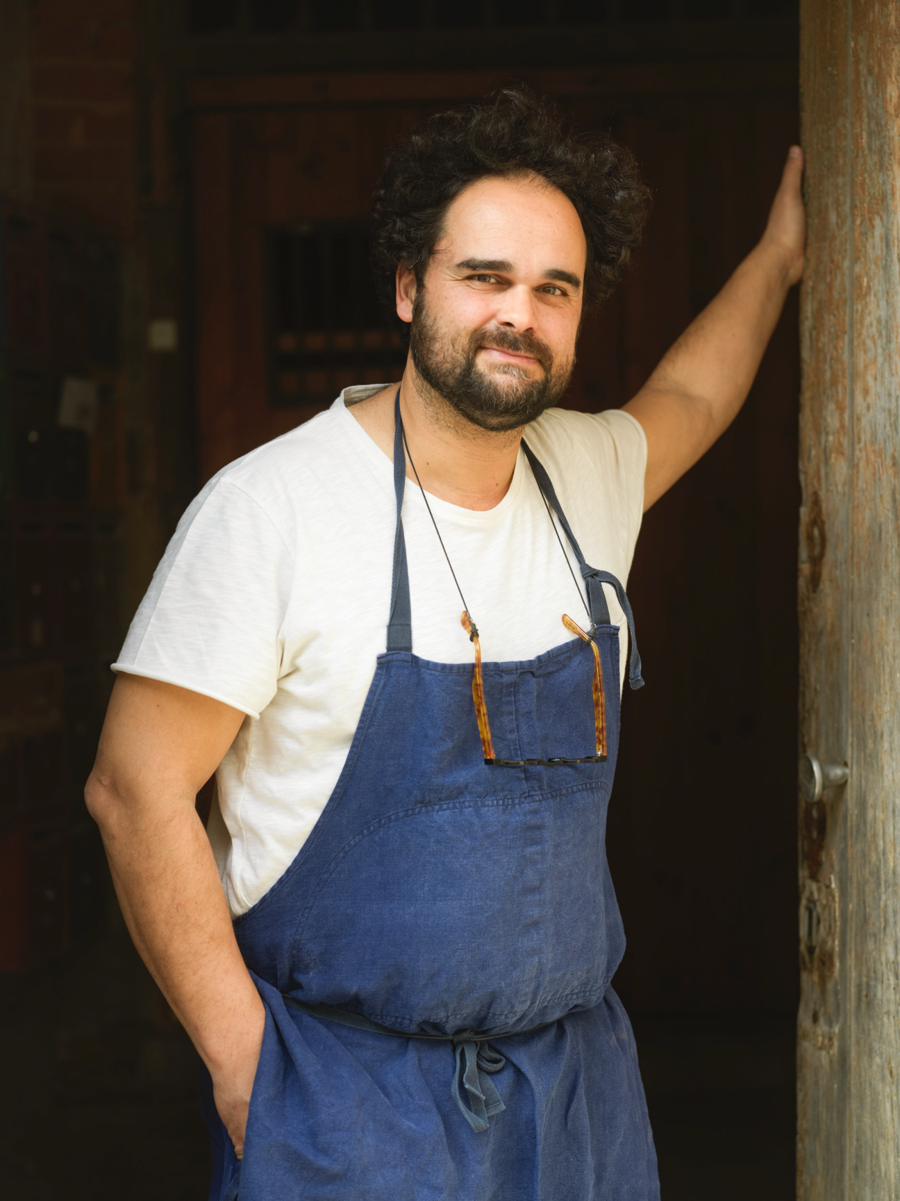 Chef in blue apron and white t-shirt smiling while leaning against wooden doorframe