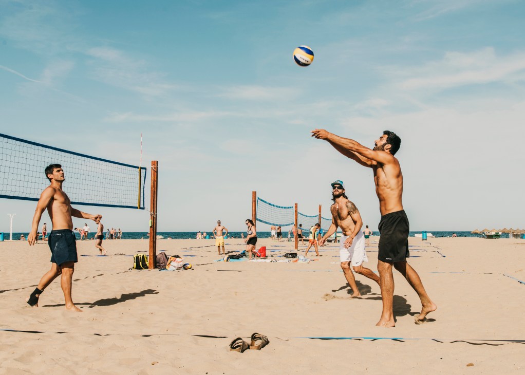 Men playing beach volleyball on golden sand with the sea and clear sky in the background.