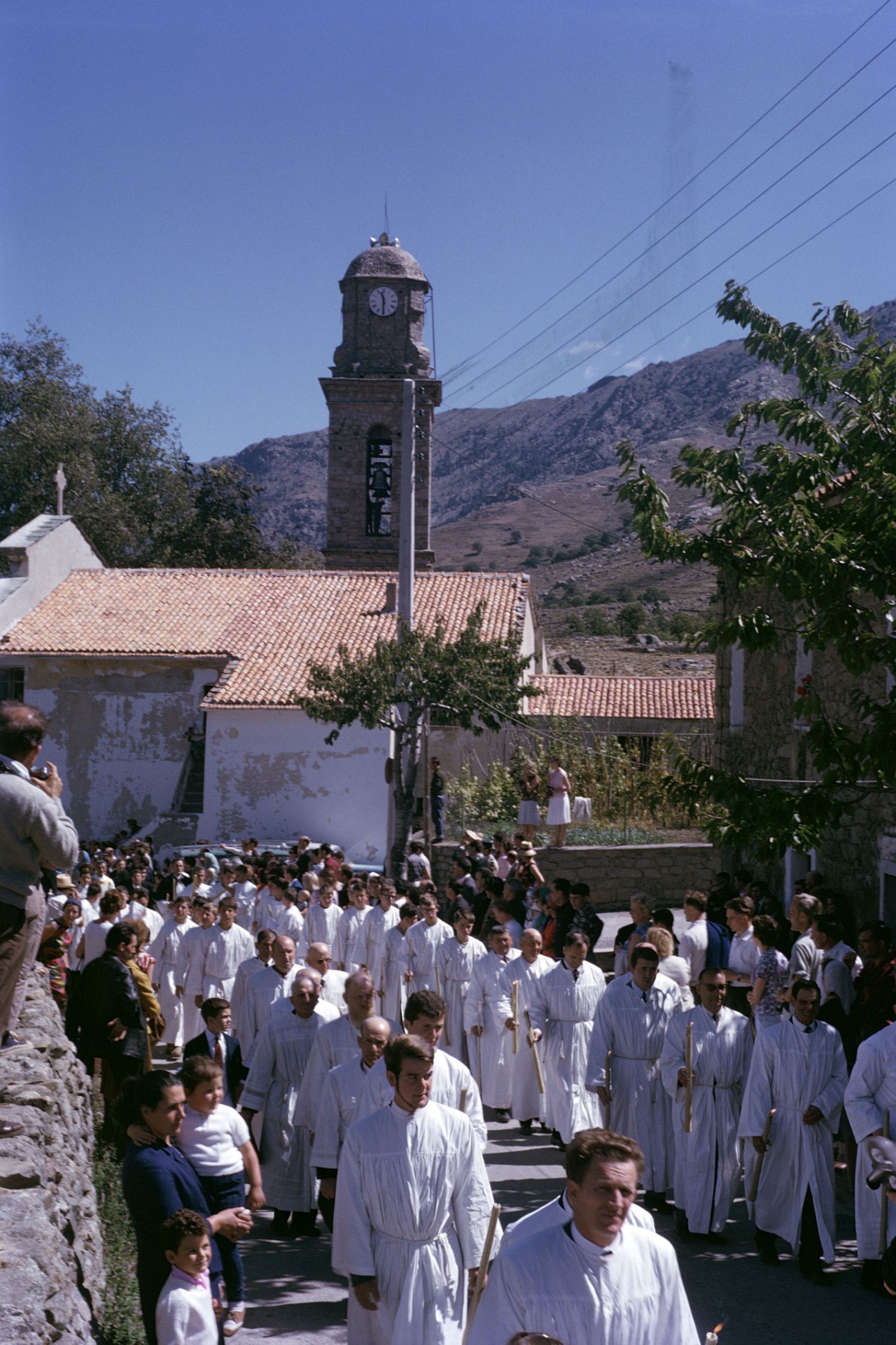 Large religious procession of people in white robes walking through Mediterranean village with bell tower