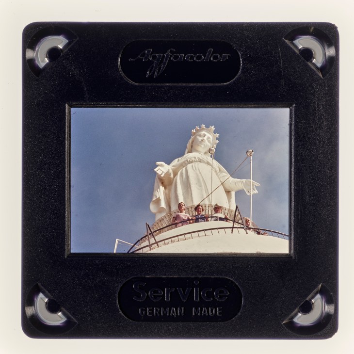 Color slide transparency showing large white religious statue with crown against blue sky, people visible at its base