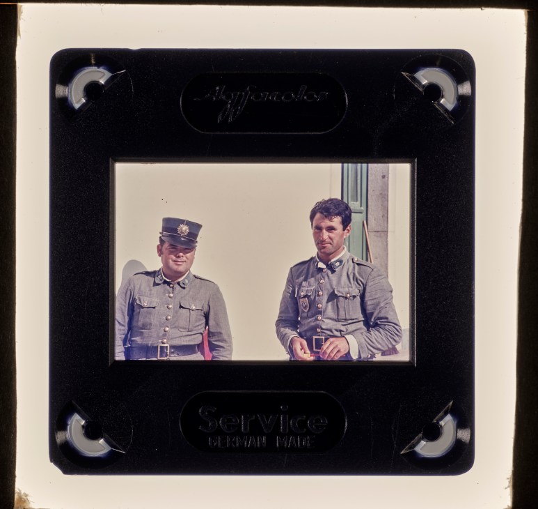 Color slide transparency showing two men in military uniforms standing outside a building, mounted in black frame