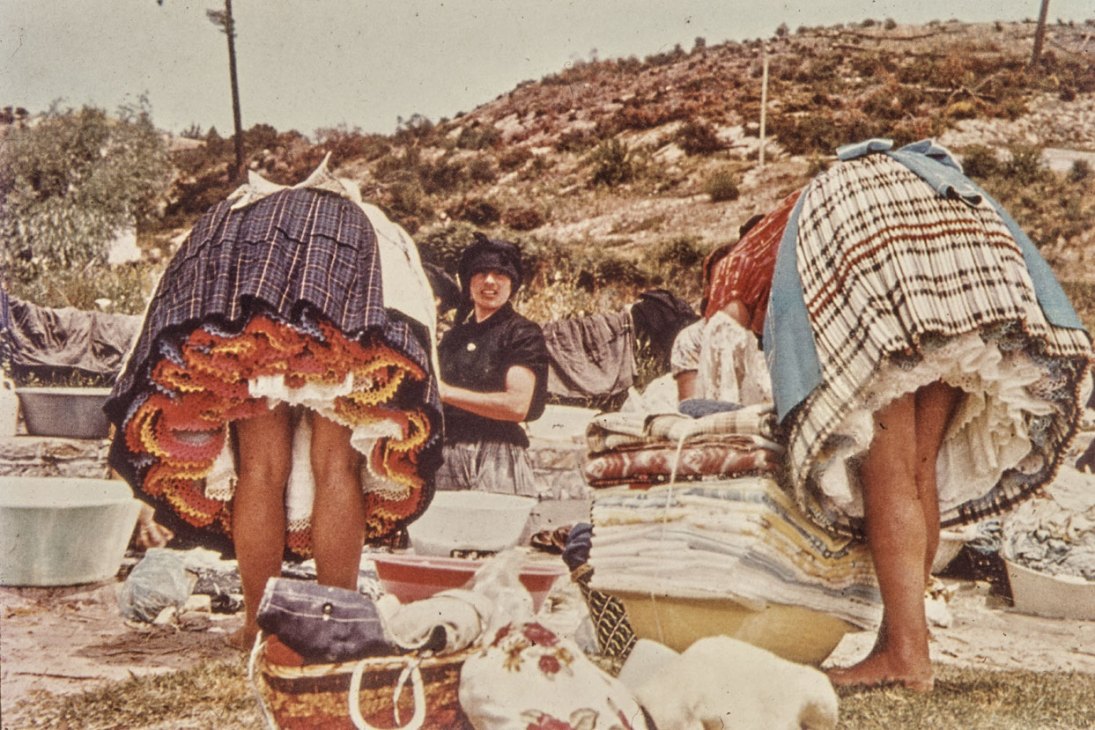 Two women in colorful traditional dresses wash clothes in basins while a man in dark clothing sits nearby