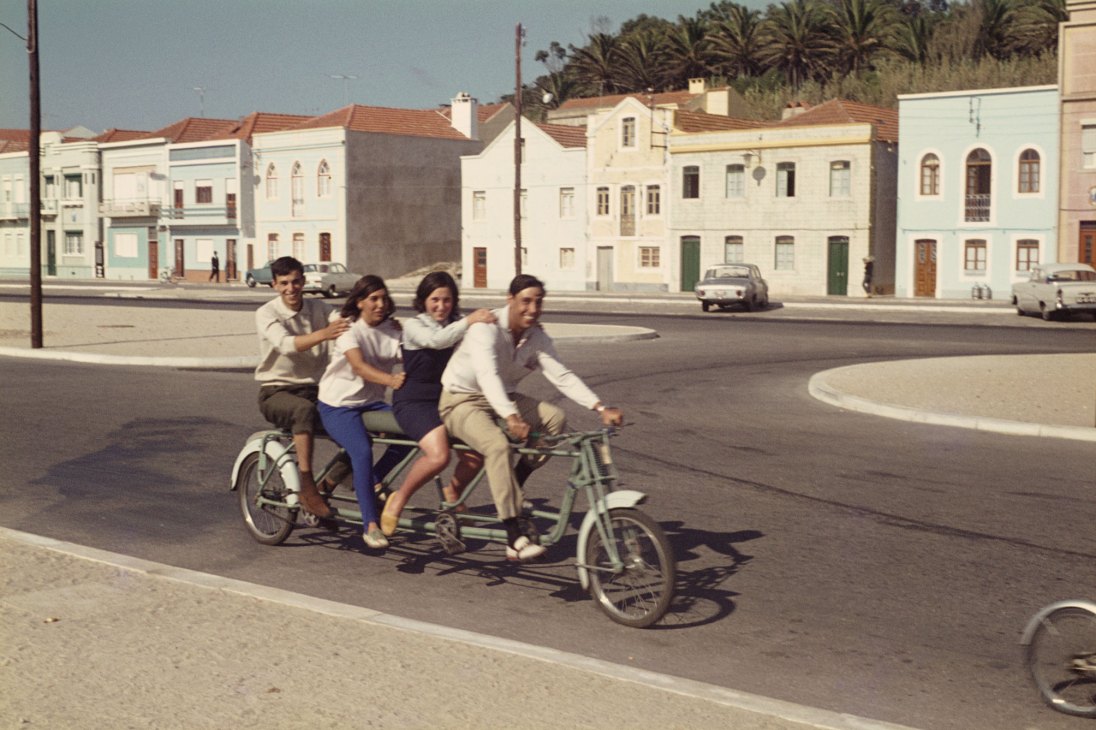 Four young people ride together on a custom four-seat bicycle through a Portuguese town square in the 1960s