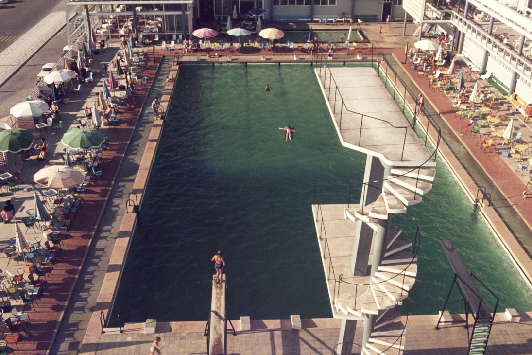 Aerial view of mid-century swimming pool with diving board, curved walkways, and sunbathers under colorful umbrellas