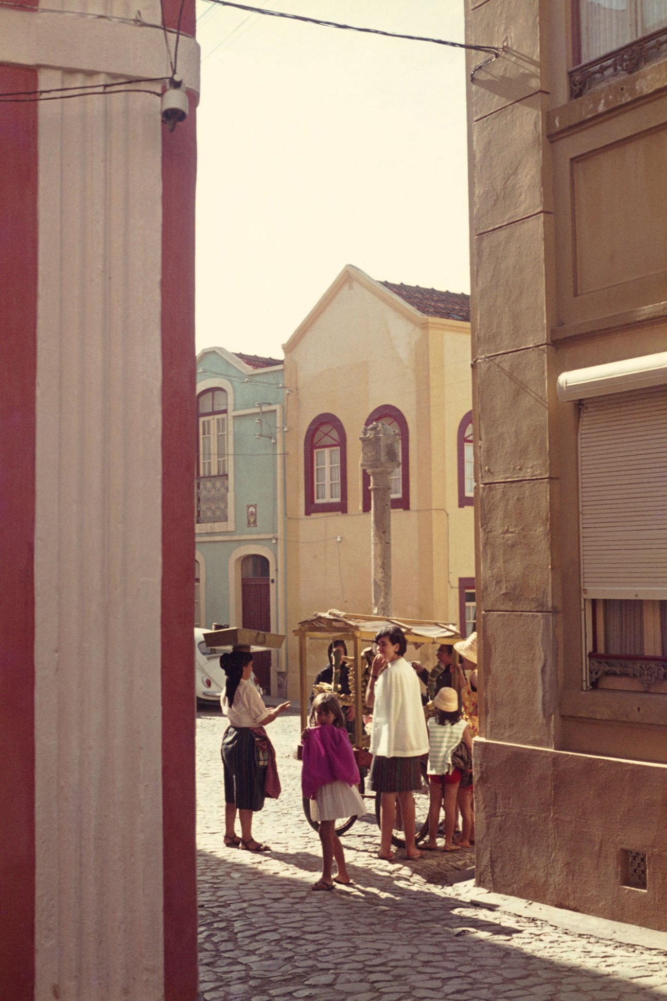 Group of people gathered around a street vendor in a Mediterranean cobblestone square with colorful buildings