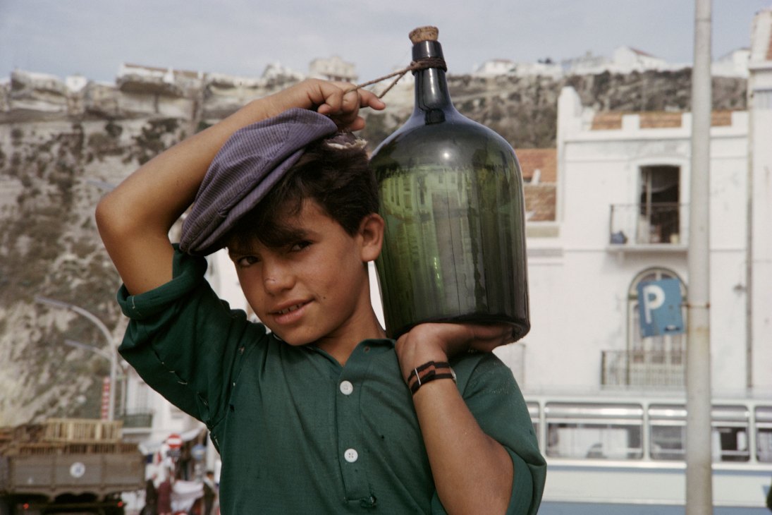 Young man in green shirt and purple head covering carrying large glass bottle in Nazaré, Portugal