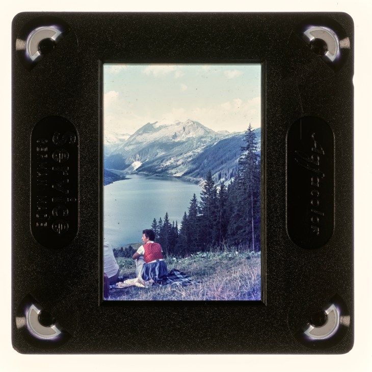 A vintage film slide showing a woman in traditional dress sitting by a mountain lake with snow-capped peaks