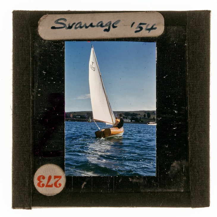 Vintage slide showing small sailboat with white sail on blue water, labeled 'Swanage '54' in damaged black frame
