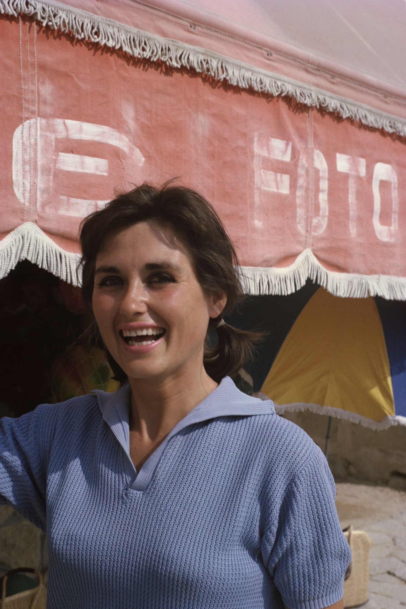 Woman in blue sweater smiling at camera while standing under pink awning with white fringe trim
