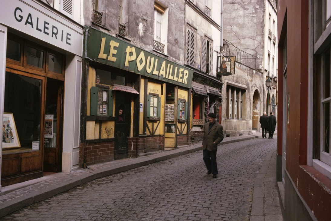 Cobblestone street scene with shops including Le Poulailler and Galerie, man walking in overcoat, postwar Paris 1953