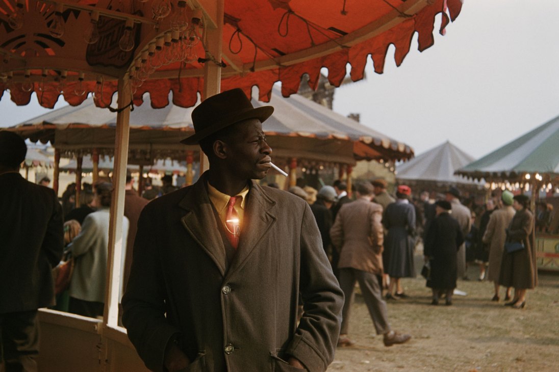 Man at a fair wearing trilby hat and tie, smoking as sunlight glints off his tiepin