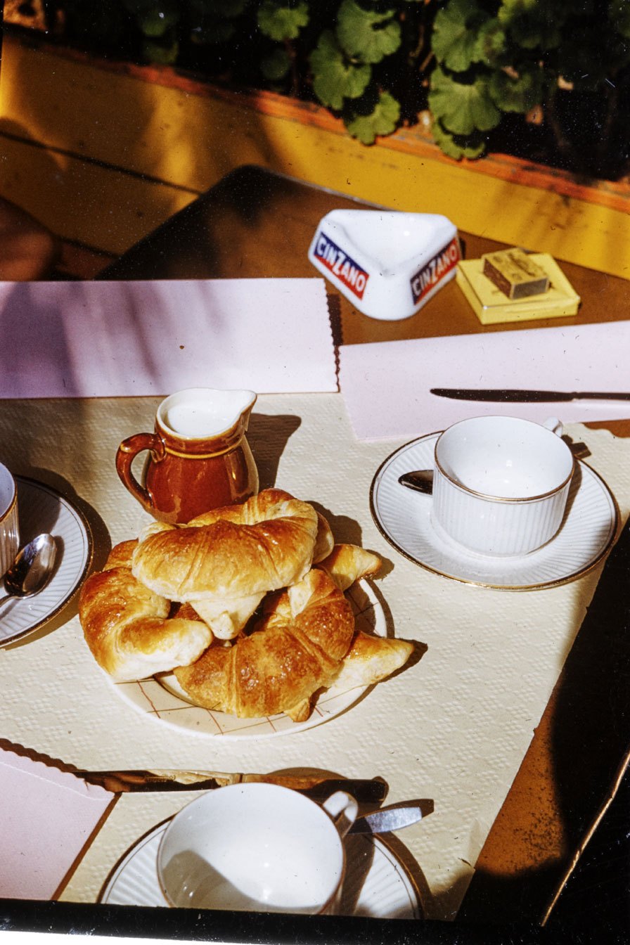 Breakfast table with croissants on wire basket, white teacups with gold trim, cream pitcher, and napkins in sunlight