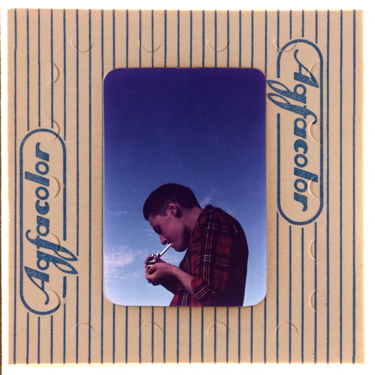 Man in plaid shirt lighting cigarette on European street in Percy Waite's mid-century color photography