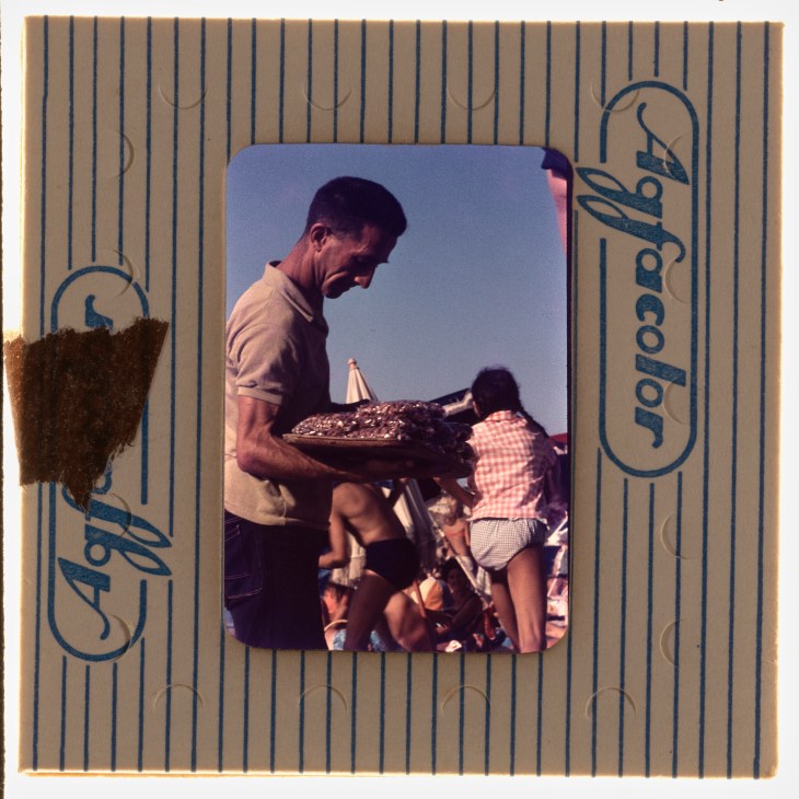 Vintage color photograph showing a man serving food at an outdoor barbecue with children seated nearby