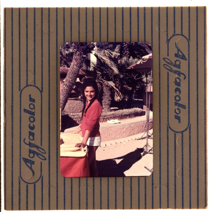 Vintage Agfacolor slide showing woman in red dress standing by poolside with palm trees in background