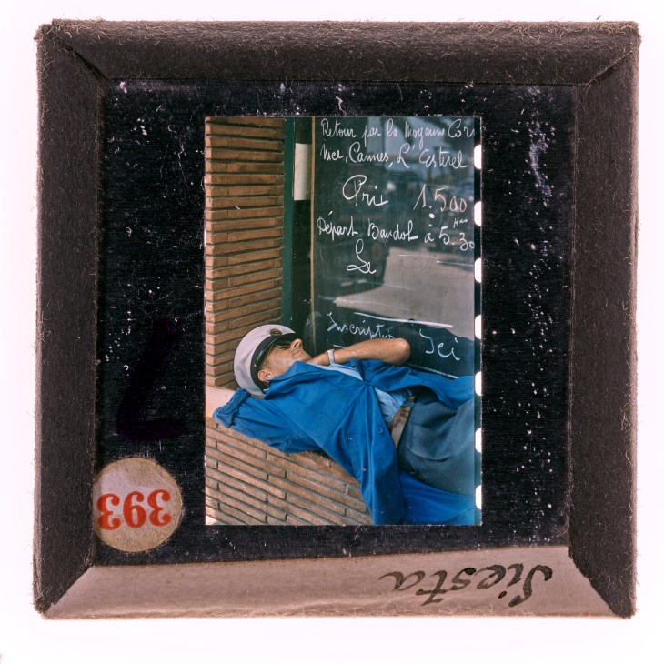 Vintage slide showing man in blue shirt writing on chalkboard in Mediterranean setting, framed in black mount