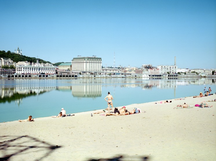 People sunbathing and swimming on a sandy beach beside calm water with city buildings reflected in background