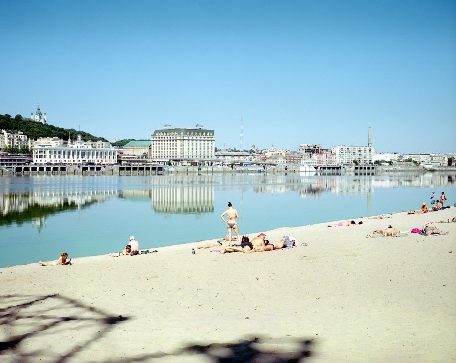 People sunbathing and swimming on a sandy beach beside calm water with city buildings reflected in background