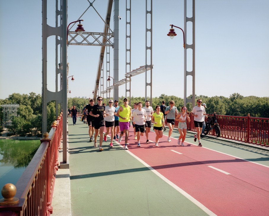 Group of runners jogging across a pedestrian bridge with red railings and white steel framework
