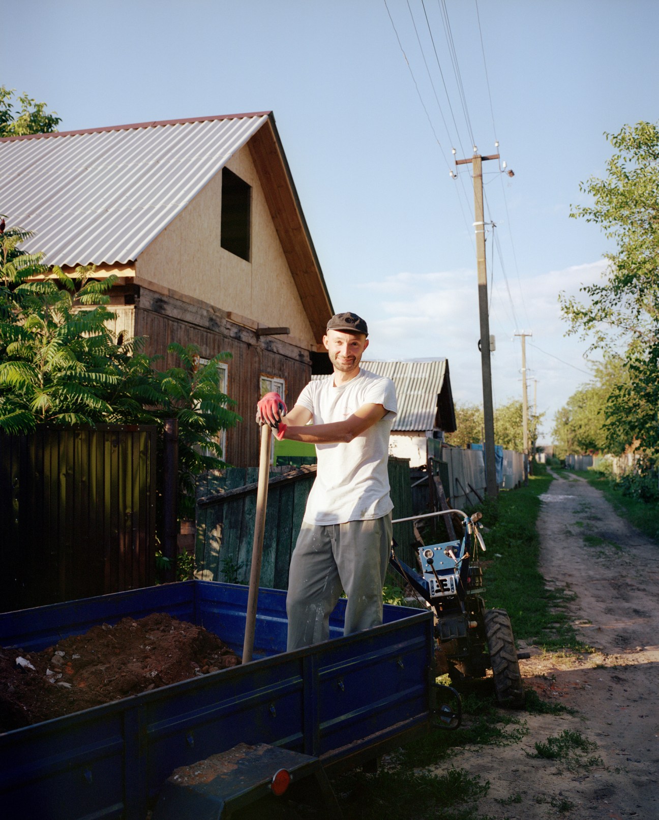 Man in white t-shirt and cap standing with shovel next to blue cart filled with dirt on rural road