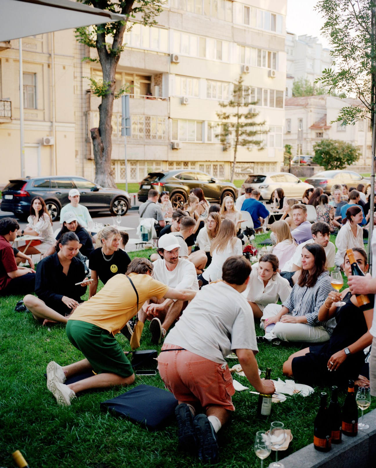 A large group of people gathered on a lawn in front of an apartment building, sitting and socializing during daytime, with parked cars visible in the background.