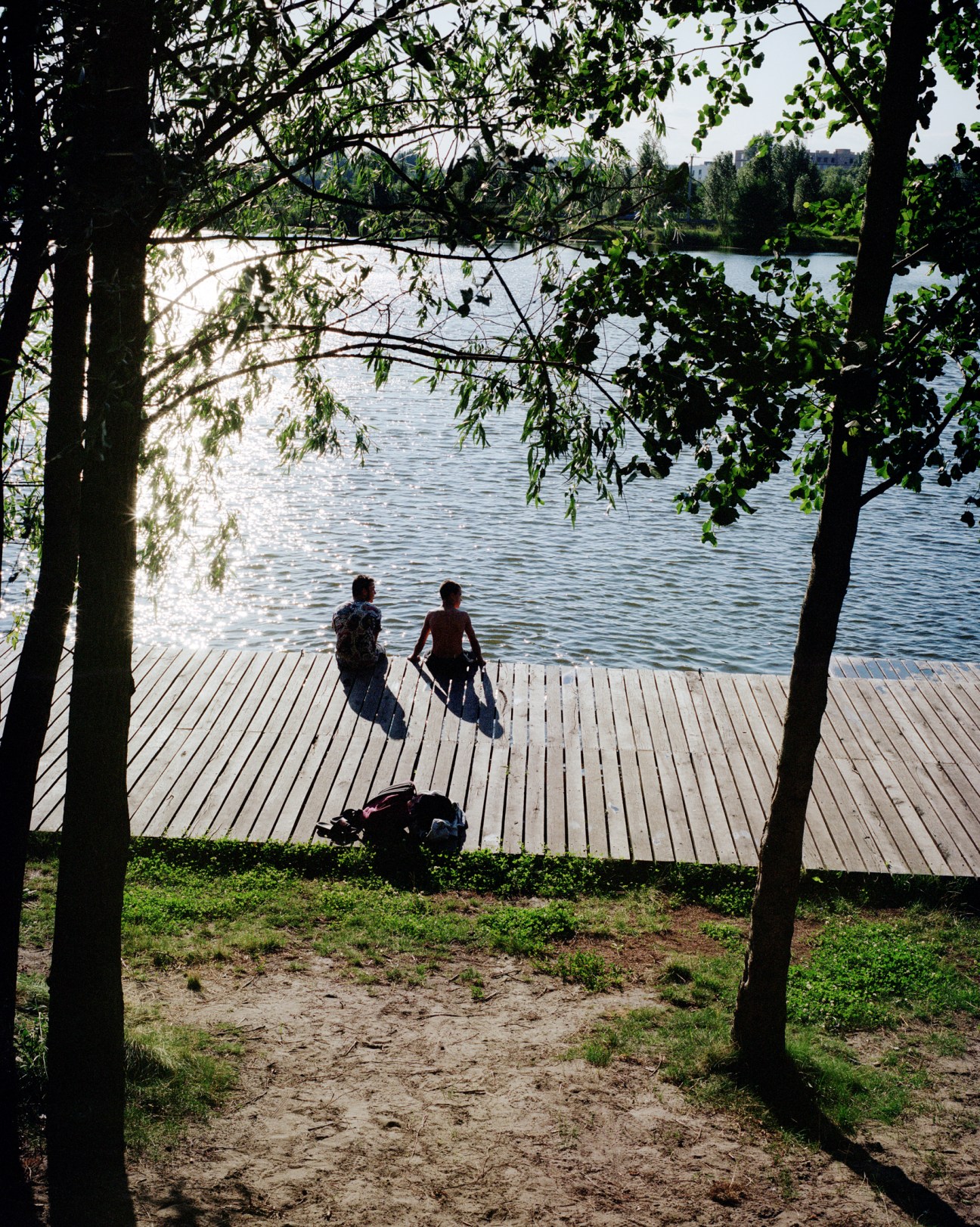 Two people sitting on wooden dock at lake in Bucha Municipal Park on summer day