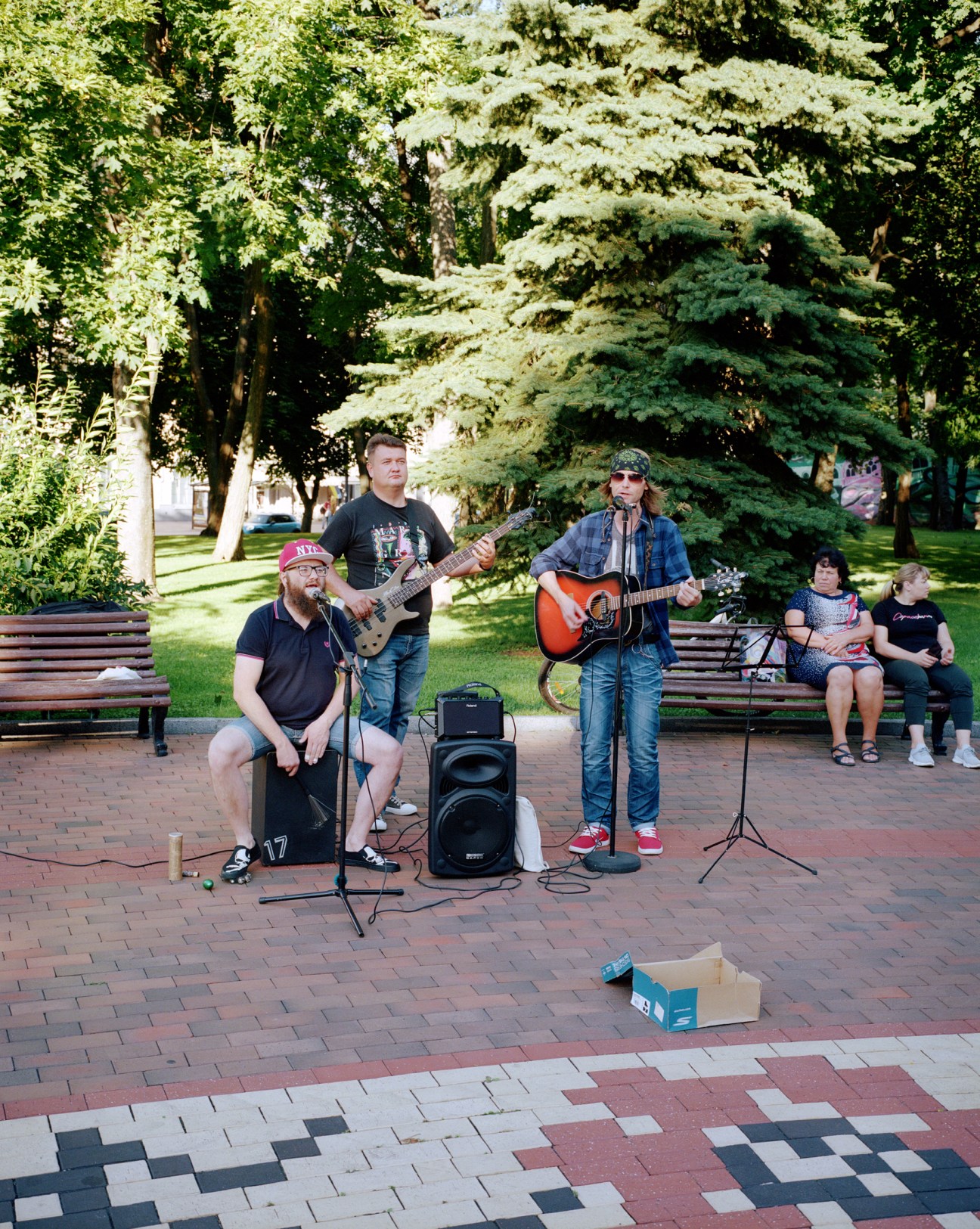 Musicians performing with guitars and microphone in a public square with benches and trees