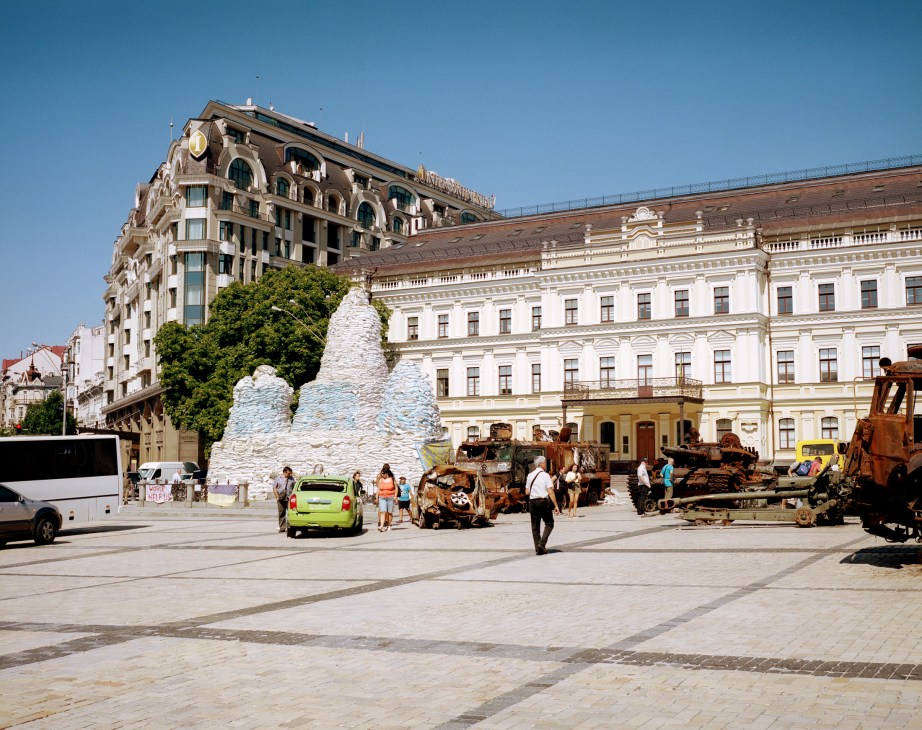 Stone monuments covered in protective white material with rusted military equipment displayed in a public square