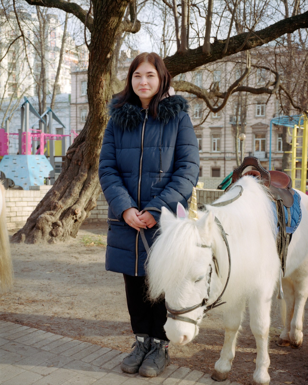 Young woman in blue winter coat standing next to white pony with saddle in park