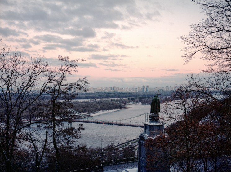Monument of Prince Volodymyr the Great overlooking Kyiv's modern skyline at dusk with river and bridge visible