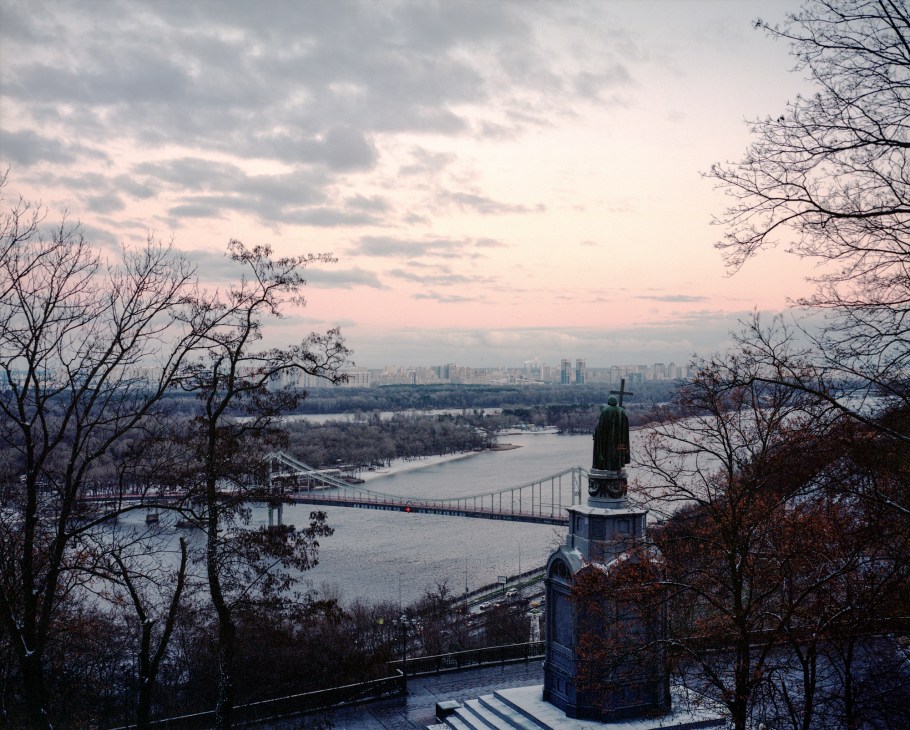 Monument of Prince Volodymyr the Great overlooking Kyiv's modern skyline at dusk with river and bridge visible