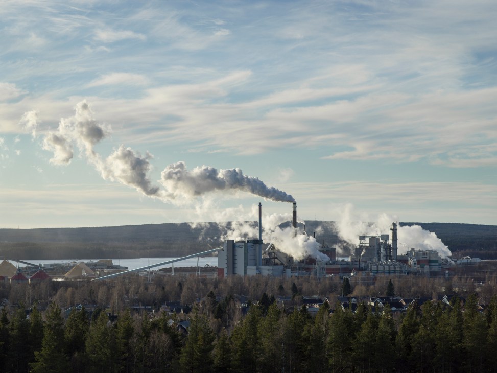 Large industrial paper mill complex with smokestacks emitting white smoke and steam against cloudy sky