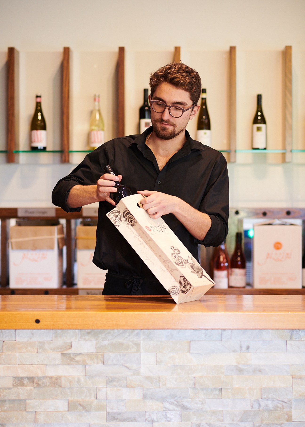 Man in black shirt opening wooden wine box at Pizzini winery with wine bottles displayed on shelves behind