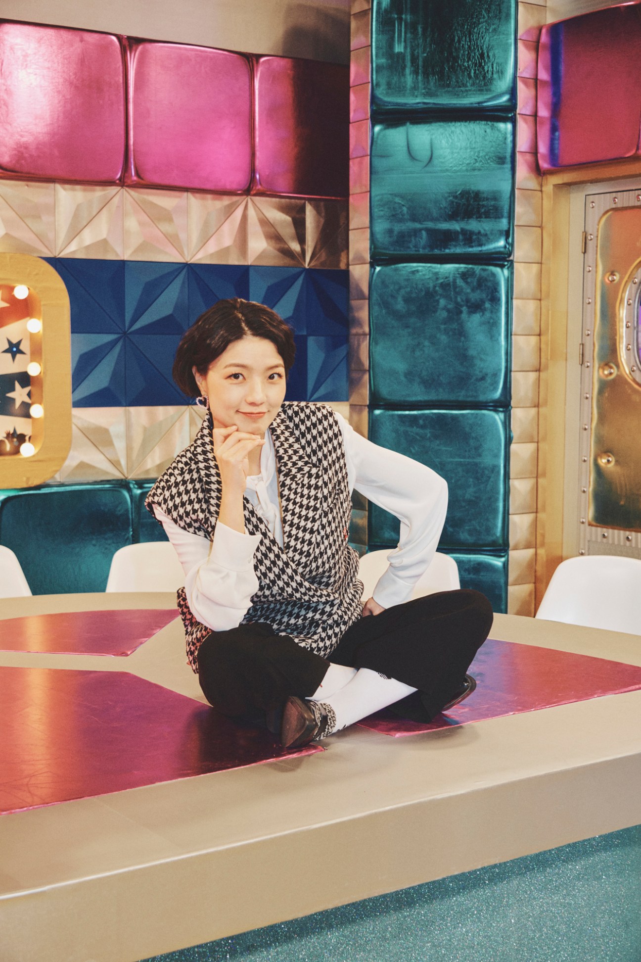 Ahn Young-mi sits cross-legged on a colorful TV studio set wearing a houndstooth vest and white shirt
