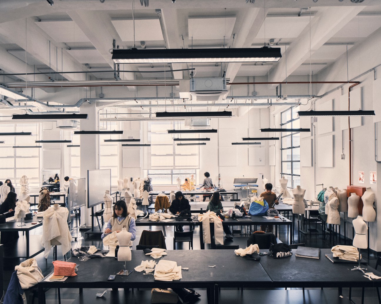Students working on pattern-making and garment construction at tables in a bright, modern fashion atelier with natural light and suspended black linear lighting fixtures
