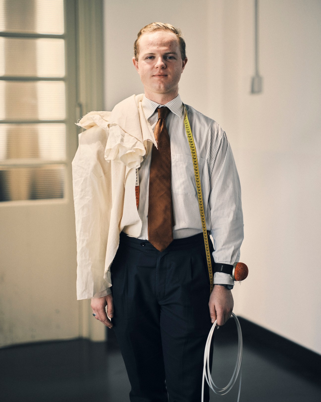 Cole Luca Heard wearing striped shirt and tie with measuring tape around neck holding white garment