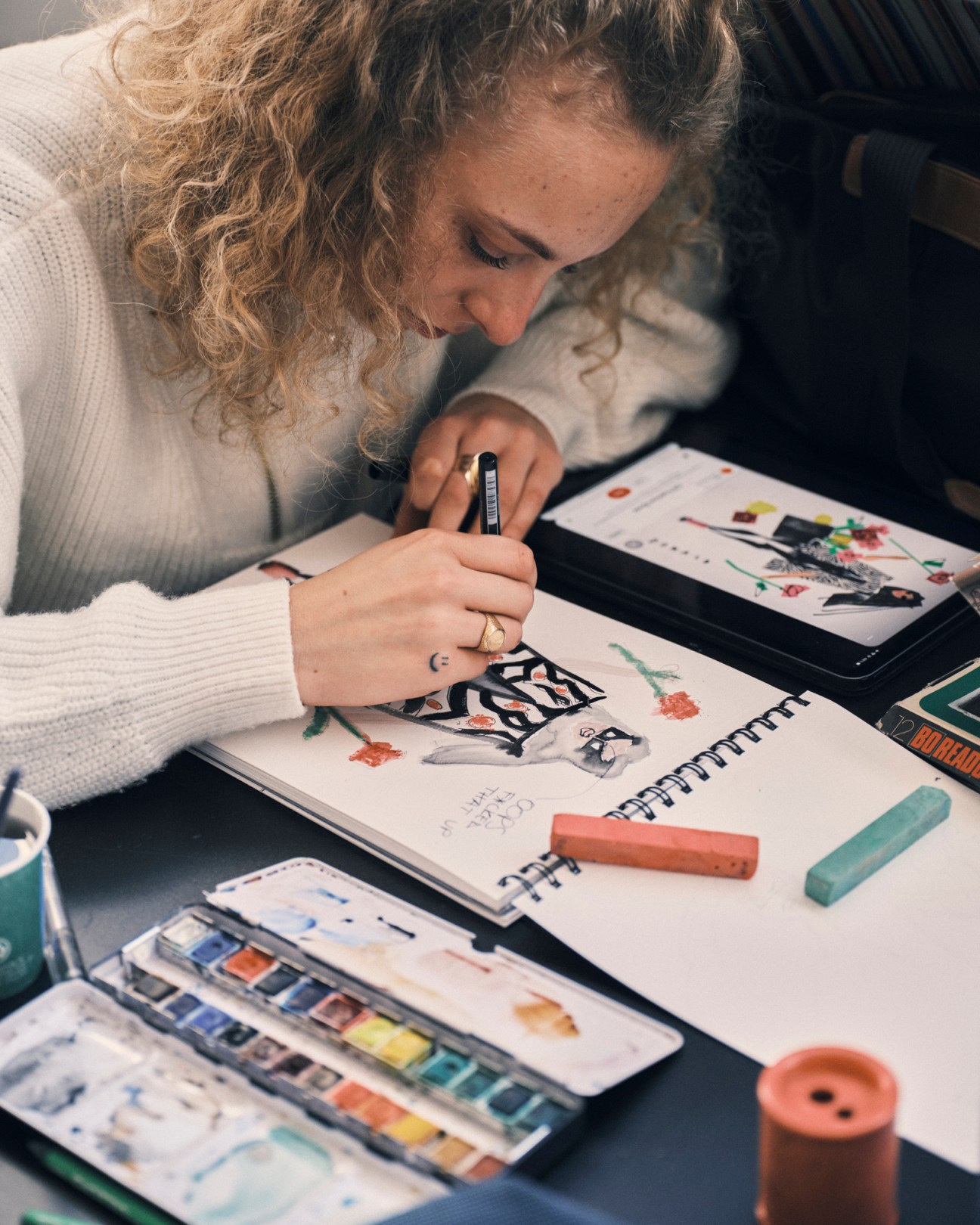 Student with curly blonde hair drawing in sketchbook at desk with watercolors, pastels, and tablet nearby
