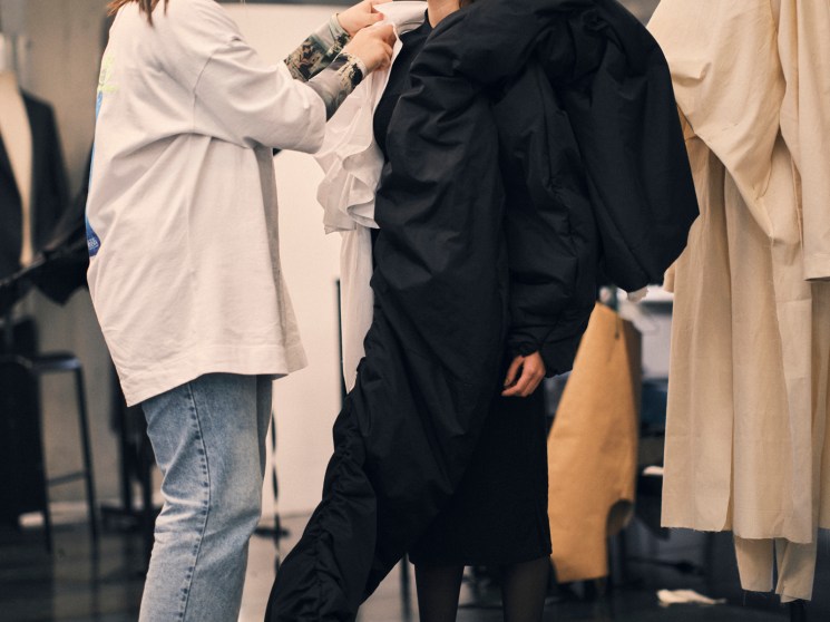 Two students in a fashion classroom, one fitting a black garment on another who is wearing protective covering