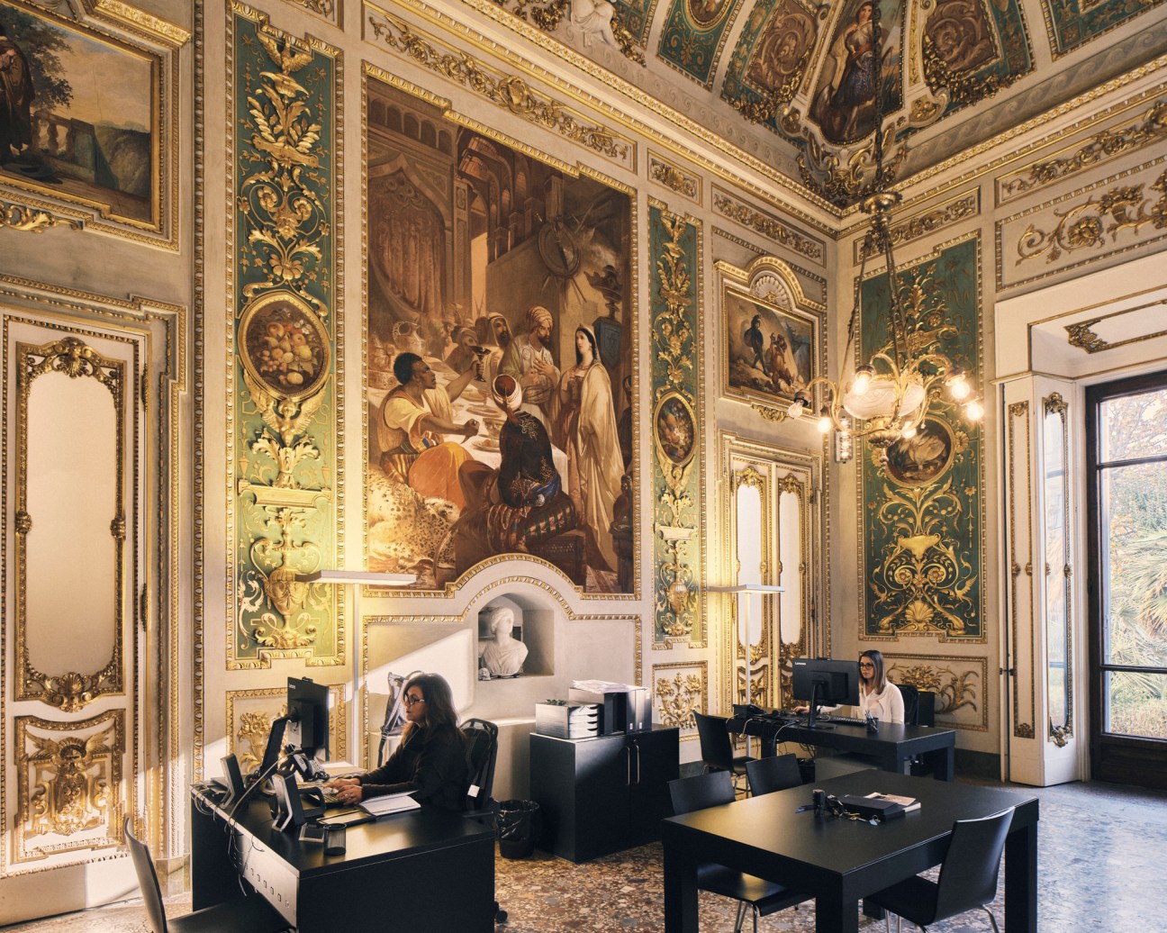 Ornate baroque office interior with frescoed walls, gilded decorations, and modern black desks where staff work