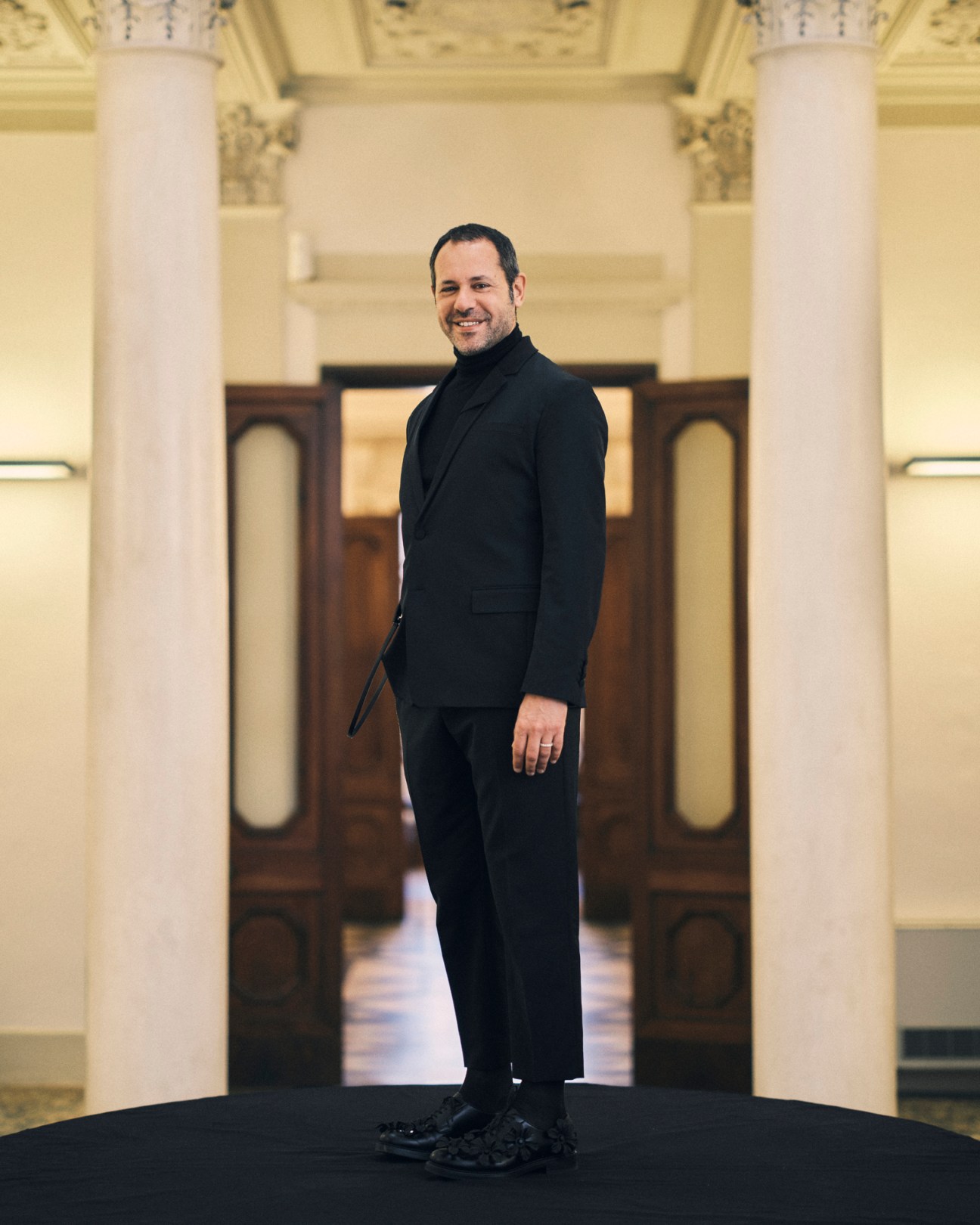 Massimiliano Giornetti in black suit standing in ornate building with classical columns and decorative ceiling