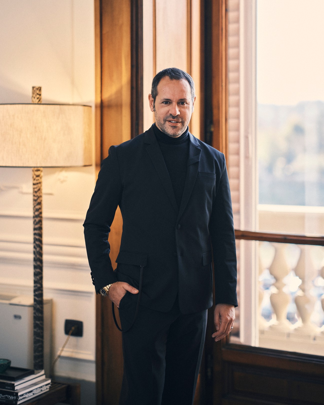 Man in dark suit jacket and black turtleneck standing by window in elegant interior