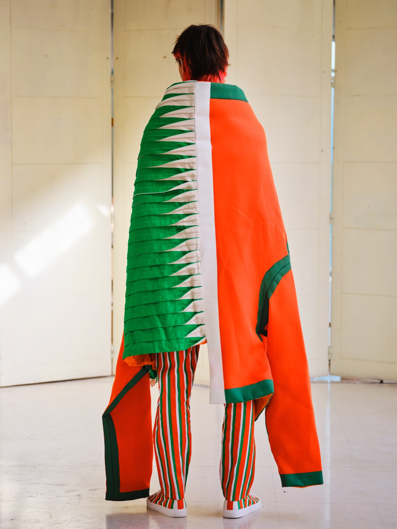 Student wearing vibrant orange and green cape-like garment with geometric patterns and matching striped pants