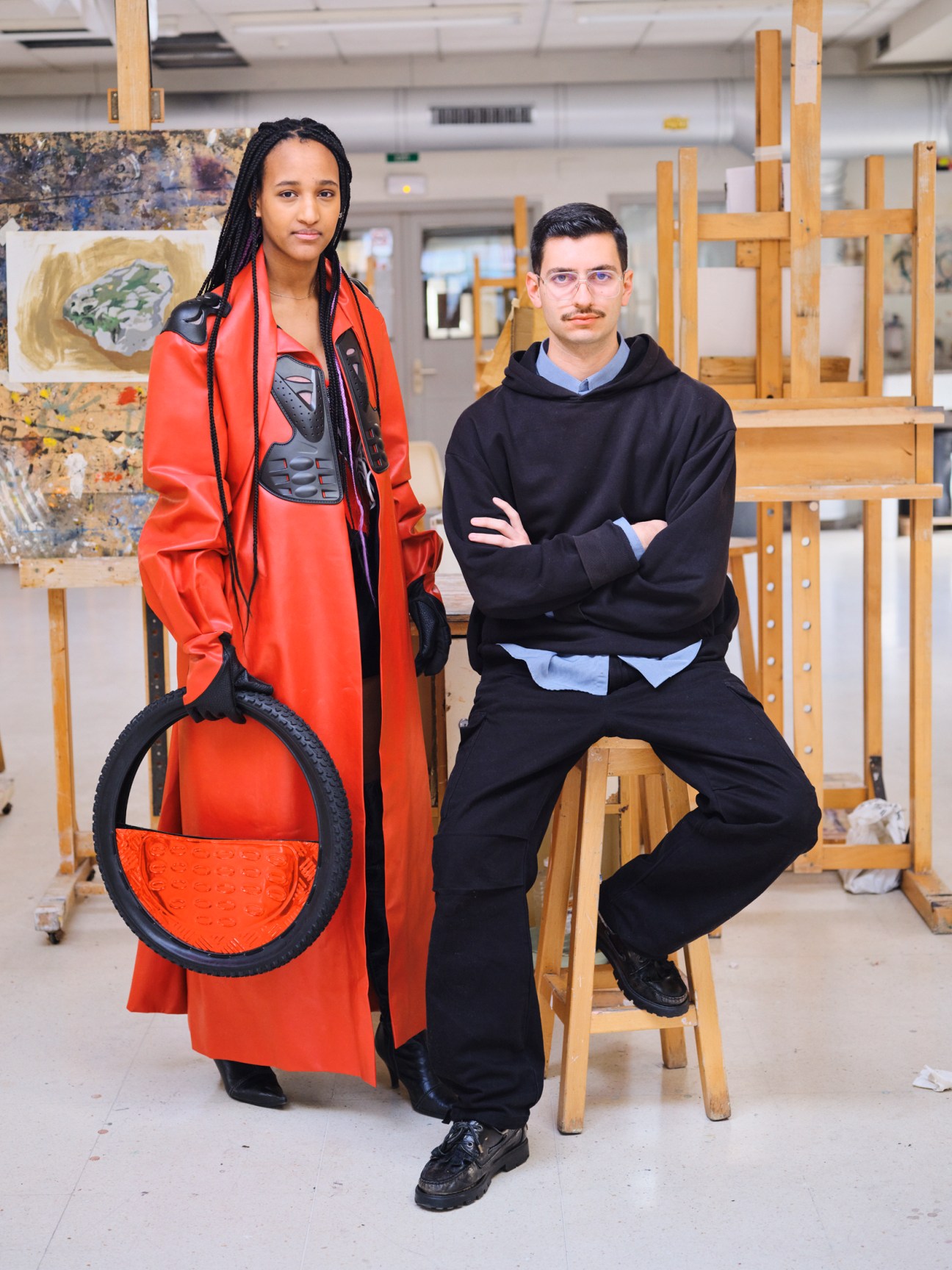 Two fashion students pose in art studio, one wearing red coat holding tire, other in black hoodie sitting on stool