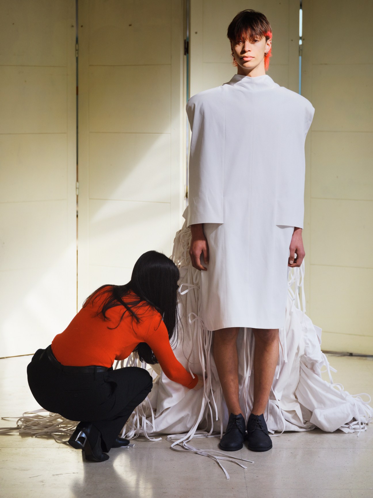 Fashion student in red top kneeling to adjust white ribbons on a minimalist white dress worn by standing model