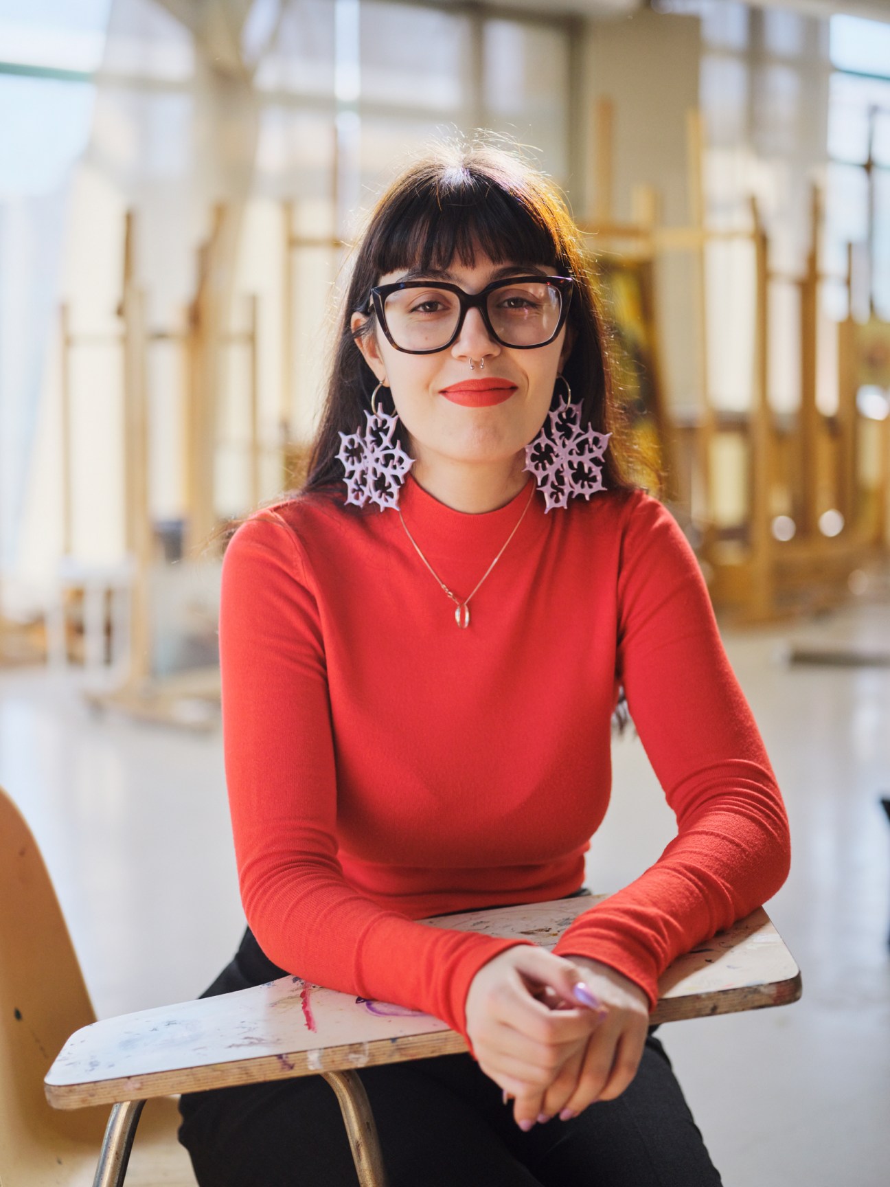 Young woman with glasses and bangs wearing red sweater and decorative earrings sitting at wooden table