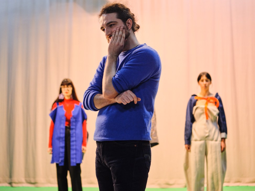Man in blue sweater covering part of his face with his hand while two people in costumes stand in background