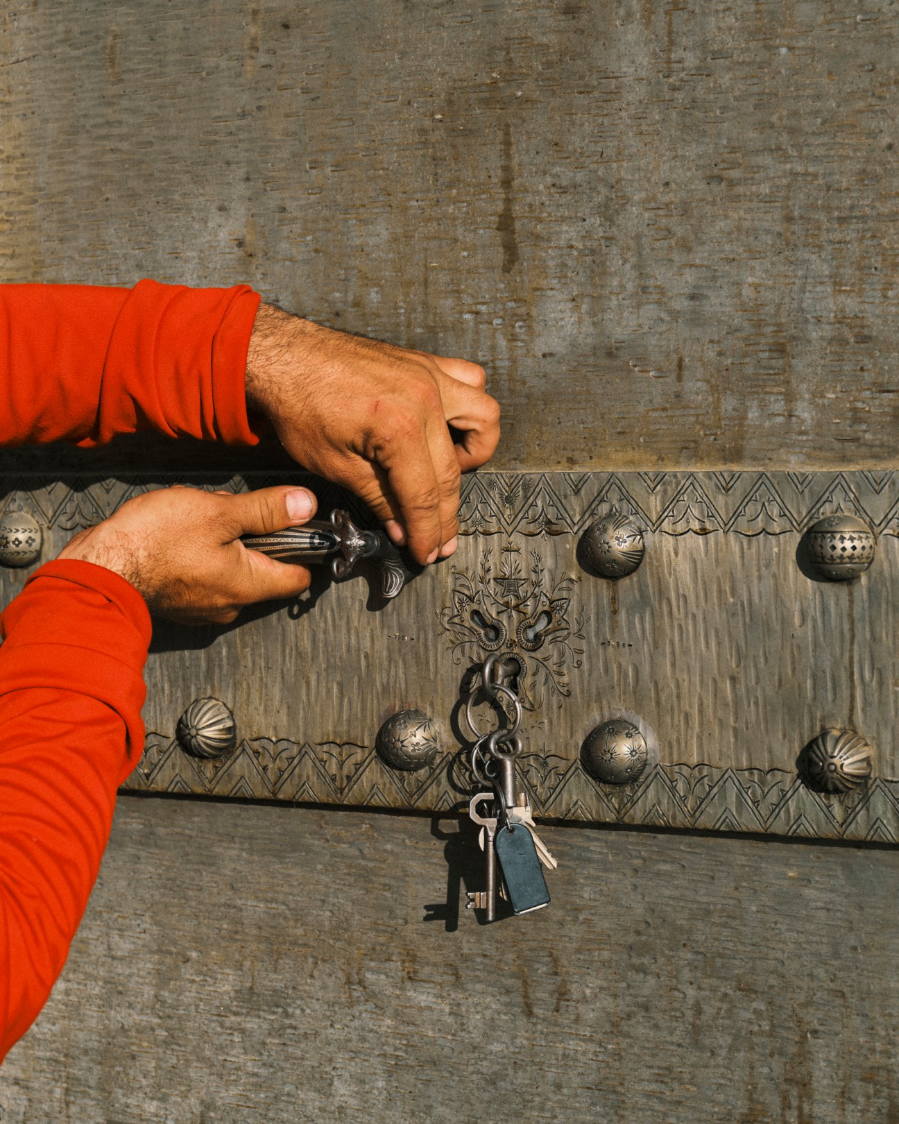 Hands in orange sleeves using key to open ornate metal door with decorative patterns and studs