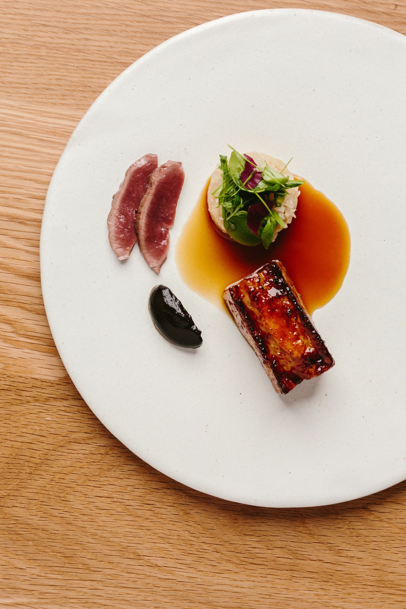 Elegantly plated dish featuring sliced seared meat, glazed rectangular protein, greens, and dark sauce on white plate