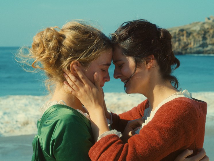 Two women touching foreheads intimately on a beach with ocean and cliffs in background