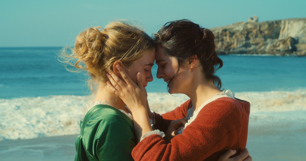 Two women touching foreheads intimately on a beach with ocean and cliffs in background